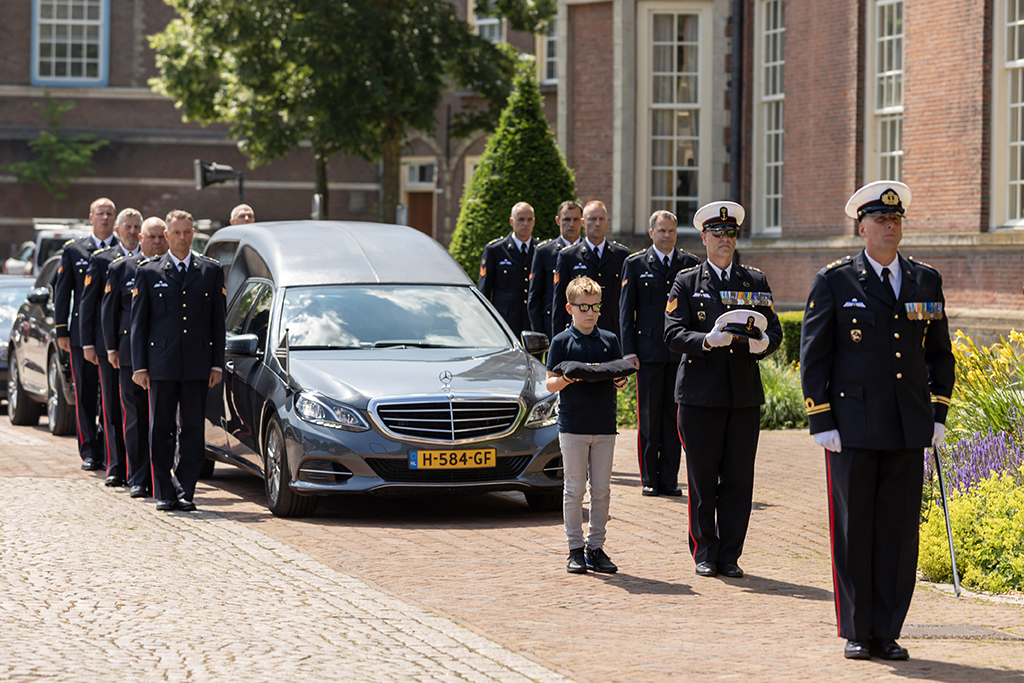 GonnieBax3 Laatste groet van twee mensen bij de kist voor het sluiten omringd door gekleurde bloemen bij het raam
